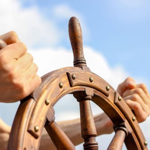person holding the wooden steering wheel of a ship with blue sky and clouds in the background