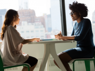 young white women and slightly older black woman at table talking by a window with city scape in the background