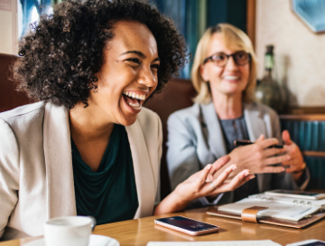 black woman laughing and white woman smiling while having coffee together