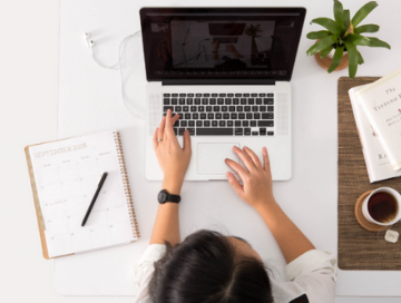 woman seated at desk that has a calendar, plant, coffee cup and laptop on it