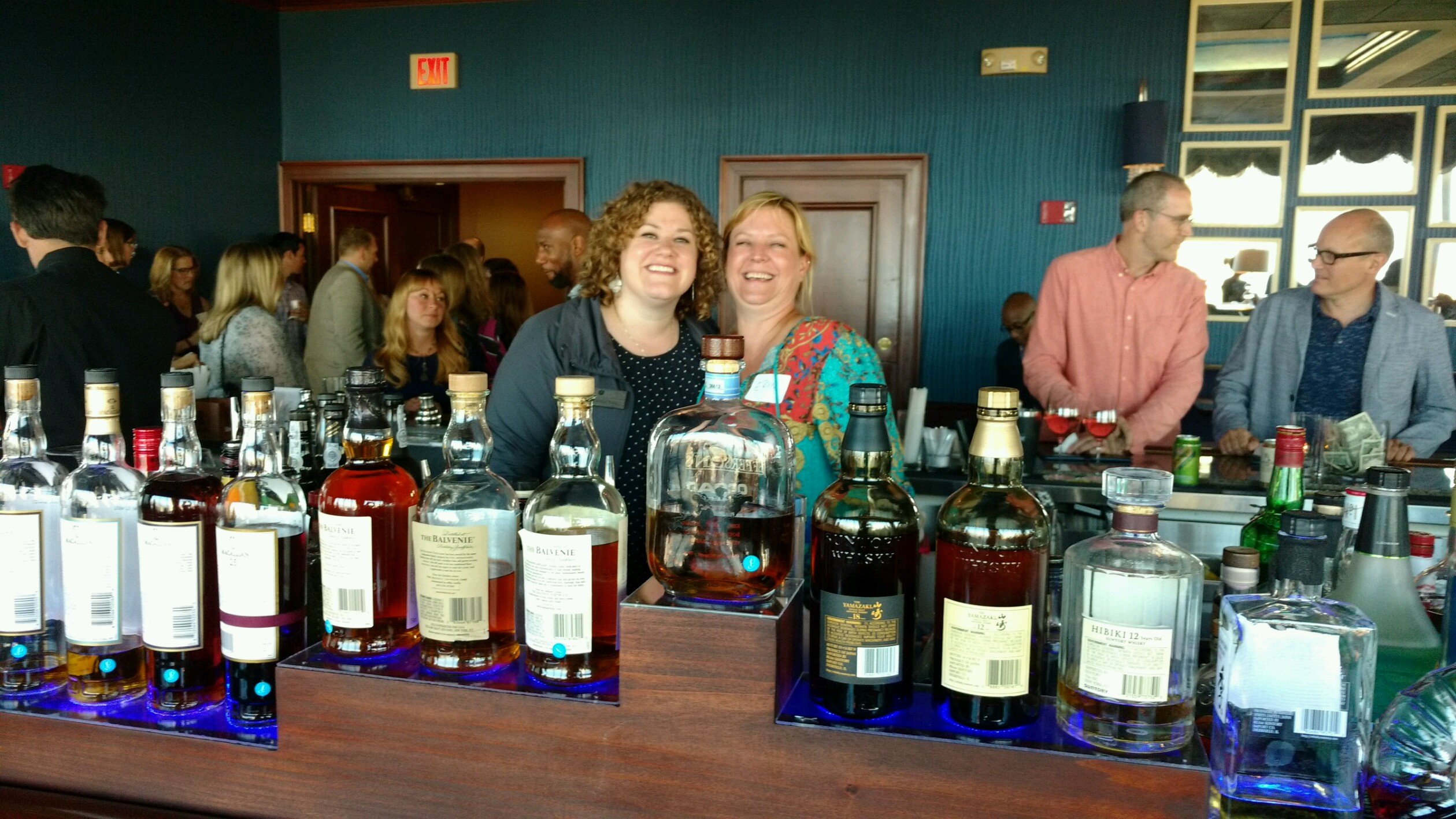 Carolyn Hahn and Erin Richardson posing while bartending. Row of liquor bottles across the bar in the foreground.