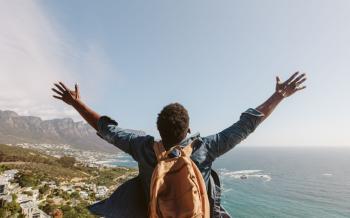 Rear view of young guy with backpack standing outdoors with arms spread open against seascape. Man enjoying the view from the top of the mountain.