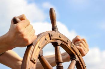 person holding the wooden steering wheel of a ship with blue sky and clouds in the background