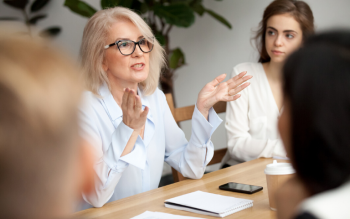 grey haired professional female talking to a conference table of people
