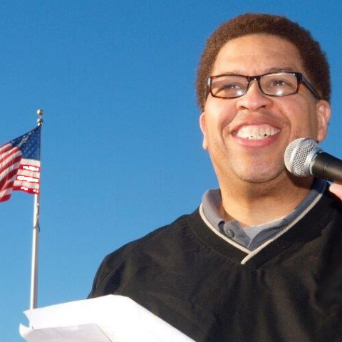 Tony Shields holding microphone presenting with American flag and blue sky in background