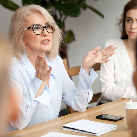 grey haired professional women talking to a group at a conference table