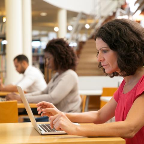 Serious focused middle aged woman working on research in public library. Adult students sitting at desks and using laptops.