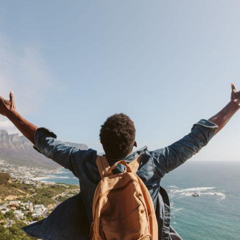 Rear view of young guy with backpack standing outdoors with arms spread open against seascape. Man enjoying the view from the top of the mountain.