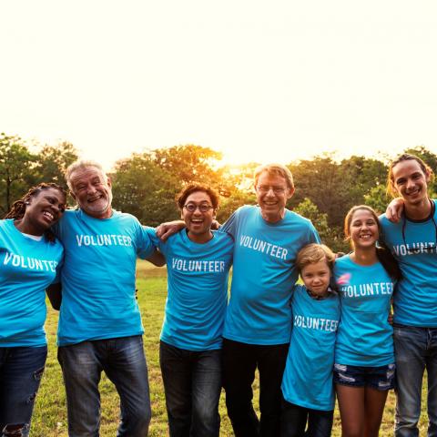 Group of people all various ages and ethnicities with arms around each other. Each wearing a blue "volunteer" shirt.