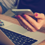 image of female hand holding mobile phone at desk working at a laptop