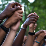 photo of many people with varying skin tones with arms raised in the air holding each others hands in unity at a protest